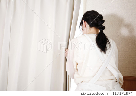 A woman in an apron looking outside through a gap in the curtains A woman in an apron looking outside through a gap in the curtains 113171974