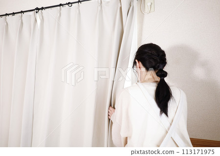 A woman in an apron looking outside through a gap in the curtains 113171975