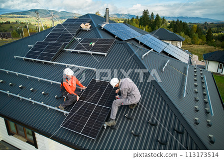 Technicians installing photovoltaic solar panels on roof of house. Men engineers in helmets building solar module system with help of hex key. Concept of alternative, renewable energy. 113173514