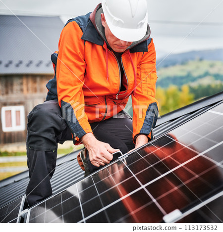 Man builder mounting photovoltaic solar panels on roof of house. Engineer in helmet installing solar module system with help of hex key. Concept of alternative, renewable energy. 113173532