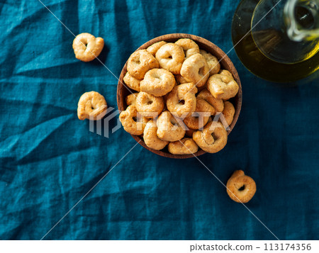 Traditional italian snack taralli or tarallini in wooden bowl over dark blue linen napkin background. Rustic shot of taralli appetizer with copy space. Top view or flat lay. 113174356