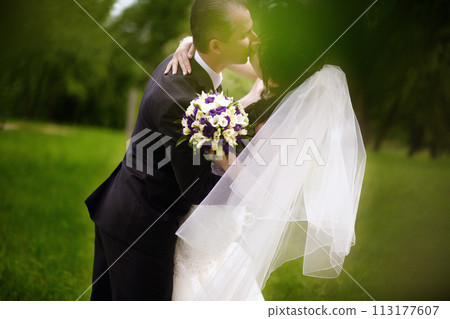A couple in a wedding dress and with a bouquet of flowers kiss in the park A couple in a wedding dress and with a bouquet of flowers kiss in the park 113177607