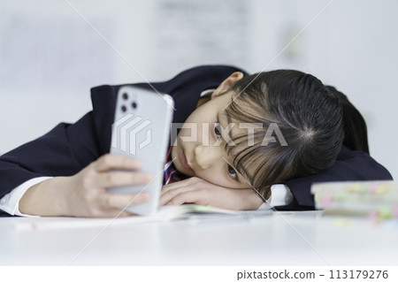 Female student lying down on the desk with a smartphone in hand 113179276