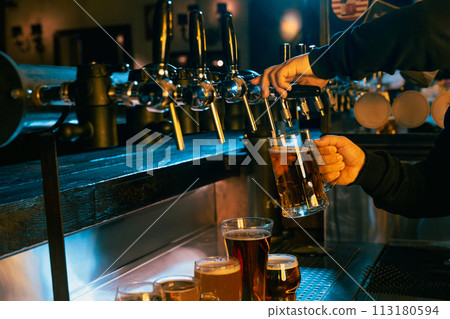 Hand drawing cold, refreshing, lager beer from tap with various other beers displayed at well-lit bar. Serving craft beer from tap. Hand drawing cold, refreshing, lager beer from tap with various other beers displayed at well-lit bar. Serving craft beer from tap. 113180594