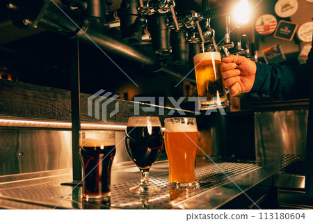 Photo of bartender's hand holds glass of light, cold, refreshing lager beer. Man Serving craft beer from tap at traditional pub. 113180604