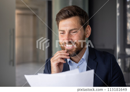 A smiling businessman in a suit examines papers, portraying confidence and professionalism in a well-lit office setting. 113181649