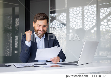 A cheerful businessman in a suit enthusiastically celebrates success while reviewing documents at his office workspace. A cheerful businessman in a suit enthusiastically celebrates success while reviewing documents at his office workspace. 113181650
