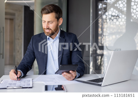 A professional man in a suit smiles as he works on paperwork in a bright modern office setting. 113181651
