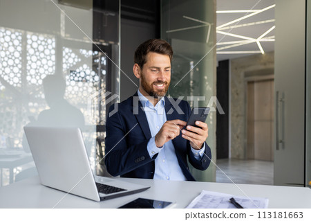 A successful and smiling young male businessman is sitting in the office at a desk in a suit and using a mobile phone. A successful and smiling young male businessman is sitting in the office at a desk in a suit and using a mobile phone. 113181663