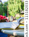 Reading glasses and open book on table in summer garden 113182033