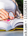 Woman's hand resting on an open book, holding reading glasses, close-up 113182034