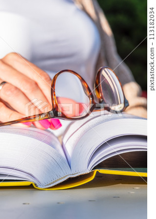 Woman's hand resting on an open book, holding reading glasses, close-up 113182034