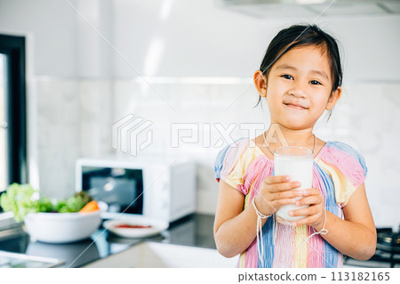 Cute Asian little girl in kitchen holds milk smiling joyfully. Portrait of daughter enjoying drink at home. Happy preschooler savoring calcium-rich liquid radiating happiness give me. 113182165
