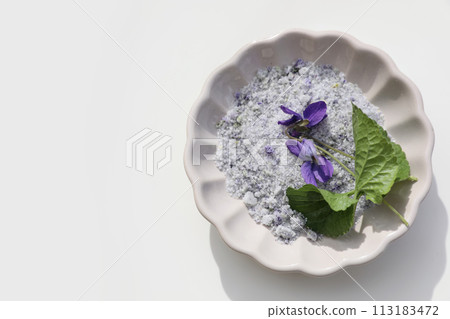 Homemade herbal sugar decorated by violet flowers and leaves. Scalloped ceramic plate isolated on white background. Viola odorata. Fragrant natural cough remedy. Food flat lay, top view. Bath salt. 113183472