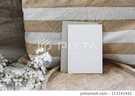 Spring still life composition. Blank greeting card mockup, craft envelope. Feminine styled photo. Floral scene. Blurred white cherry tree blossoms on wooden tray. Yellow striped linen cushion. Front 113183482