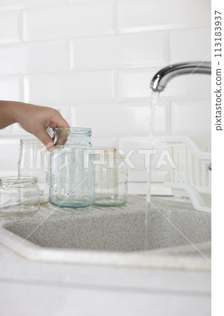 Vertical. Glass sorting. In the kitchen sink, a woman under the tap washes a glass jar to be sent for recycling. A woman sorts garbage to be sent for recycling and reuse. Eco activism concept. Vertical. Glass sorting. In the kitchen sink, a woman under the tap washes a glass jar to be sent for recycling. A woman sorts garbage to be sent for recycling and reuse. Eco activism concept. 113183937