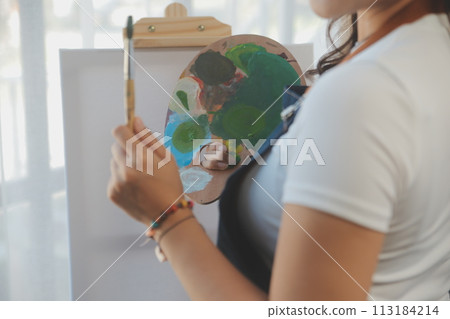 Cropped image of female artist standing in front of an easel and dipping brush into color palette Cropped image of female artist standing in front of an easel and dipping brush into color palette 113184214