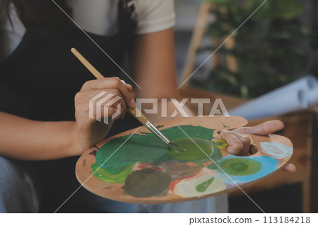 Cropped image of female artist standing in front of an easel and dipping brush into color palette 113184218