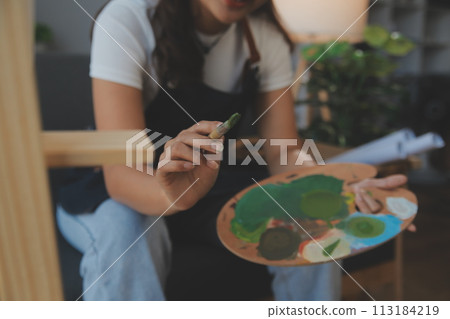 Cropped image of female artist standing in front of an easel and dipping brush into color palette 113184219
