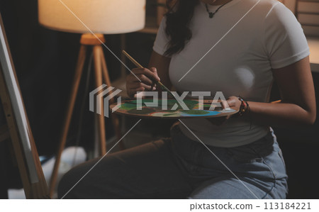 Cropped image of female artist standing in front of an easel and dipping brush into color palette Cropped image of female artist standing in front of an easel and dipping brush into color palette 113184221