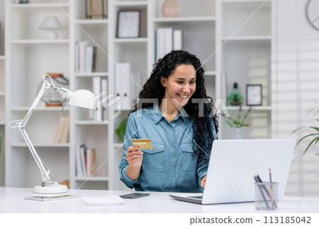 Smiling Hispanic woman conducting online transactions from her comfortable home office, showcasing remote work and financial management. Smiling Hispanic woman conducting online transactions from her comfortable home office, showcasing remote work and financial management. 113185042
