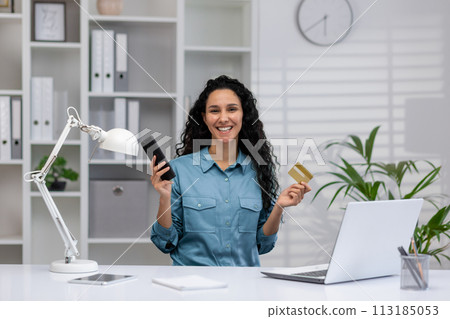 Happy Hispanic businesswoman with smartphone and credit card, working from a cozy modern home office, smiling confidently. 113185053