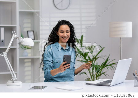 Cheerful Latina businesswoman using smartphone and holding credit card, working from a well-organized home office with laptop and green plants around. 113185062