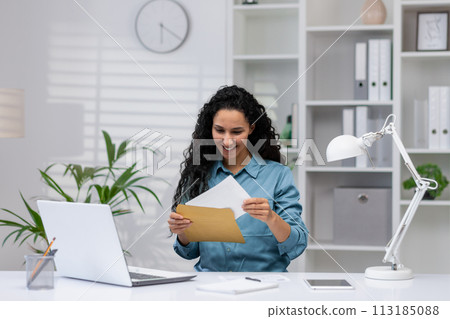 A cheerful Hispanic woman works from a well-organized home office, exuding professionalism and contentment as she handles documents. 113185088