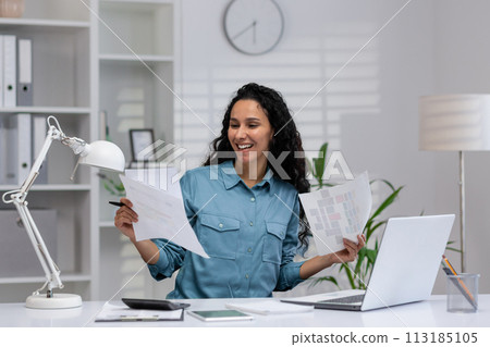 Cheerful Hispanic woman reviewing papers with a smile in her well-organized home office setting, embodying professional comfort and productivity. 113185105