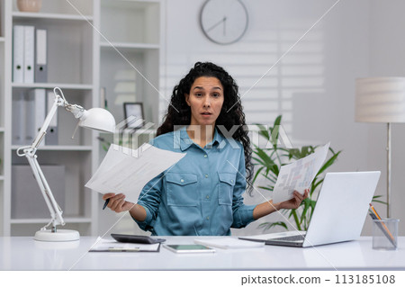 A Latina businesswoman looks confused while multitasking at her home office, surrounded by paperwork and a laptop. 113185108