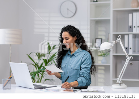A cheerful Hispanic woman manages paperwork in her modern home office, exuding professionalism and positivity. 113185135