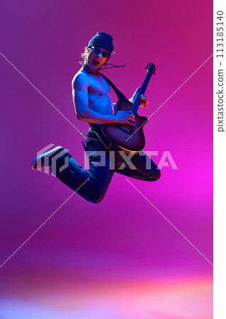 Energetic young shirtless man in sunglasses,rocket playing guitar against pink background in neon light 113185140