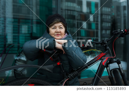 Healthy pensioner woman sitting on a bench with a bicycle on the street in winter 113185893