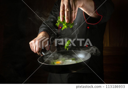 Cooking fried eggs in the pub kitchen. The chef adds fresh herbs to a steaming hot frying pan. Low key breakfast cooking concept with space for recipe. Grande cuisine Cooking fried eggs in the pub kitchen. The chef adds fresh herbs to a steaming hot frying pan. Low key breakfast cooking concept with space for recipe. Grande cuisine 113186933