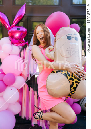 Vertical portrait of happy excited young woman in summer dress posing holding funny colorful festive foil helium balloons standing on background of country house, looking away with joyful expression. 113187949