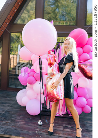 Vertical portrait of beautiful tanned blonde female in summer dress posing holding various colorful festive helium balloons standing at country house building exterior, smiling looking at camera. Vertical portrait of beautiful tanned blonde female in summer dress posing holding various colorful festive helium balloons standing at country house building exterior, smiling looking at camera. 113187950