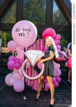 Vertical portrait of sensual blonde female in summer elegant dress posing holding various colorful festive foil helium balloons standing on background of country house building, sexy looking down. 113187951
