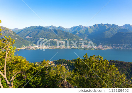 A beautiful mountain range with a lake in the foreground. The lake is calm and the mountains are covered in trees. The sky is clear and the sun is shining brightly. The scene is peaceful and serene 113189402