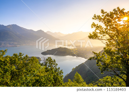 A beautiful mountain range with a lake in the foreground. The lake is calm and the mountains are covered in trees. The sky is clear and the sun is shining brightly. The scene is peaceful and serene 113189403