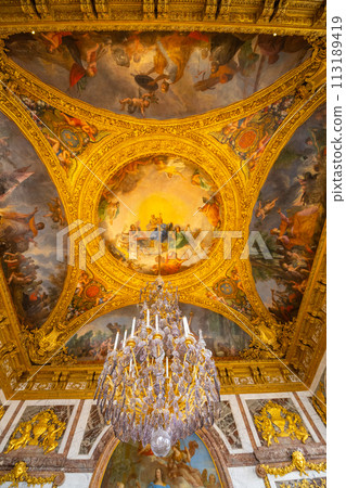 A grand chandelier hangs from the ornate ceiling frescoes of Chateau Versailles, showcasing baroque artistry. Versailles near Paris, France 113189419