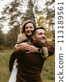 Adult couple having fun in the autumn park outdoors, happy man is carrying a woman on his back in the woods 113189561