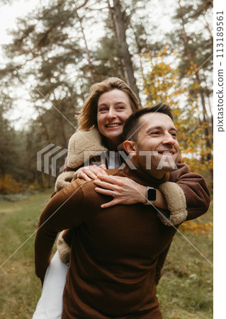 Adult couple having fun in the autumn park outdoors, happy man is carrying a woman on his back in the woods 113189561