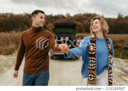 Smiling couple holding hands, strolling down country road with car on the background, happy man and woman on a roadtrip 113189567