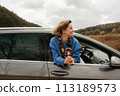 A woman leans out of the car window during road trip in autumn, the wind blows her hair 113189573