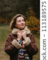 Happy caucasian woman in a brown jacket is standing in the autumn woods and cheerfully looking up 113189575