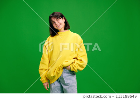 Portrait of young brunette woman with short hair posing in yellow sweatshirt and looking at camera against vibrant green studio background. Portrait of young brunette woman with short hair posing in yellow sweatshirt and looking at camera against vibrant green studio background. 113189649