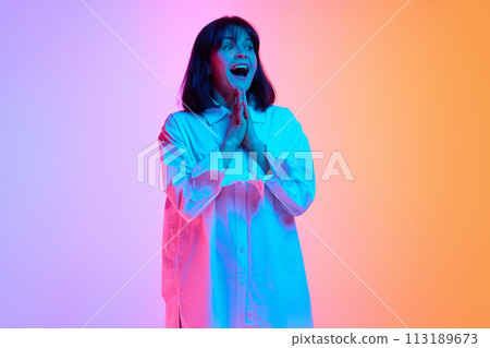 Woman with short haircut in oversized shirt with playful expression looking at camera in neon light against gradient studio background. Woman with short haircut in oversized shirt with playful expression looking at camera in neon light against gradient studio background. 113189673