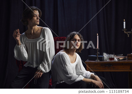 Portrait of two handsome men in white blouses sitting at room in vintage setting with candle, quill, inkwell and antique books. 113189734
