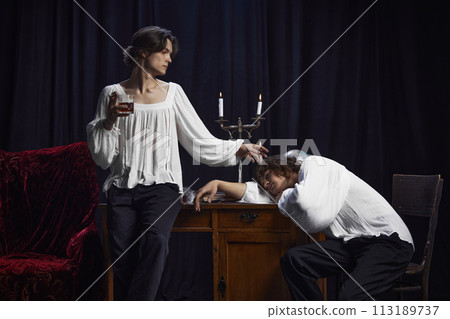 Portrait of man stands holding drink while another rests his head on desk in period-styled room with vintage furniture. Portrait of man stands holding drink while another rests his head on desk in period-styled room with vintage furniture. 113189737