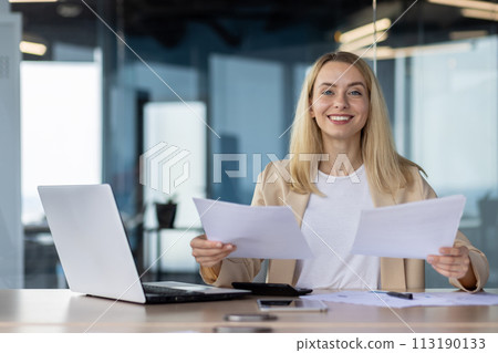 Smiling businesswoman sitting at her desk with laptop, reviewing papers in a well-lit modern office. Smiling businesswoman sitting at her desk with laptop, reviewing papers in a well-lit modern office. 113190133